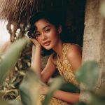 Woman in yellow dress by hut surrounded by foliage.