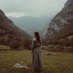 Woman in dress stands in scenic mountain landscape.
