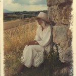 Woman in white sits by stone wall in field.