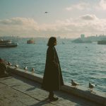 Person standing by waterfront with seagulls and boats.
