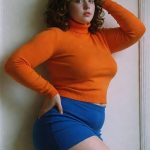 Woman in orange sweater and blue skirt posing